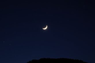 A serene night sky with the crescent moon and its companions, including Jupiter and Saturn, shining brightly amidst a canvas of stars, with a silhouette of trees or a hill in the foreground.