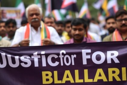 A photo of a protest in Bengaluru with a banner that reads "Justice for Ballari" in the foreground, and a blurred image of a politician in the background.