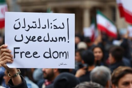 A photo of an Iranian protester holding a sign that reads "Freedom" in English and Persian, with a blurred background of a crowded street and a hint of the Iranian flag.