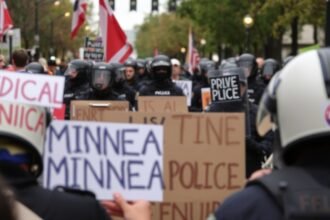 A photo of a protest in Minneapolis with a background of federal agents in riot gear, highlighting the tension between law enforcement and the community.