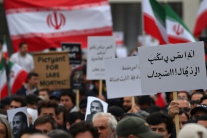 A photo of a protest in Iran with a crowd of people holding signs and banners, with a subtle background of the Iranian flag